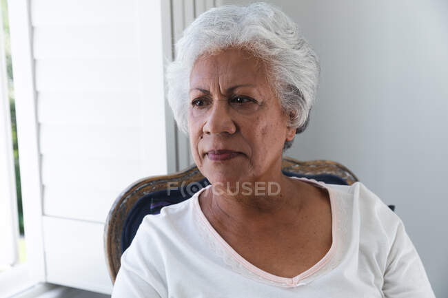 Porträt einer attraktiven Afroamerikanerin im Ruhestand mit kurzen weißen Haaren, die an einem sonnigen Sommertag zu Hause auf einem Stuhl vor einem Fenster mit weißen Fensterläden sitzt und sich während der Coronavirus-Covid19-Pandemie selbst isoliert — Stockfoto