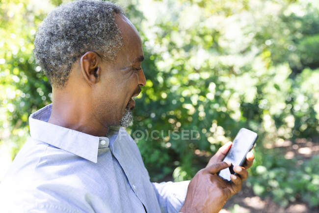 Щасливий високопоставлений афроамериканець, який тішиться своїм виходом на пенсію, у сонячному саду, спілкується по мобільному телефону і посміхається — стокове фото