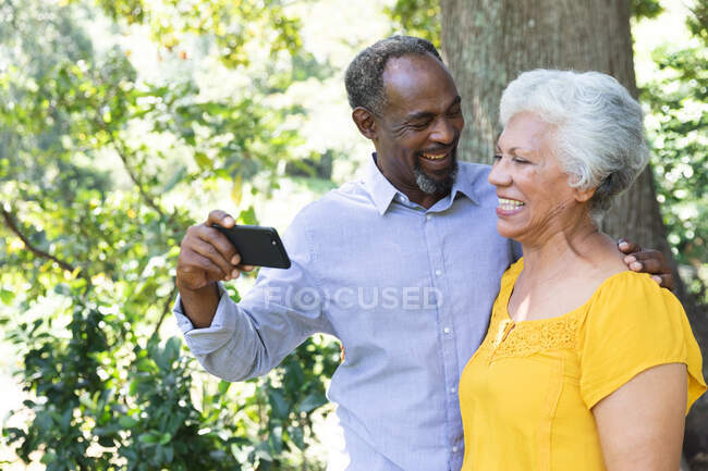 Una coppia afroamericana anziana che si gode la pensione, in piedi in un giardino al sole che abbraccia e distoglie lo sguardo sorridente — Foto stock