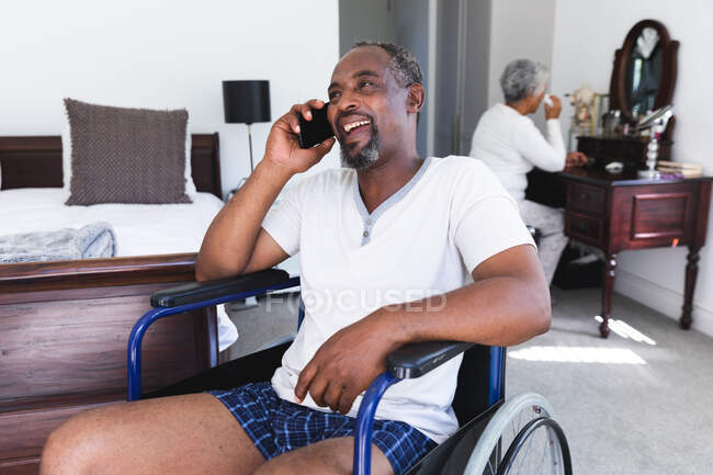 Ein älteres afroamerikanisches Rentnerehepaar zu Hause im Schlafzimmer, der Mann im Rollstuhl in Unterwäsche, der mit einem Smartphone spricht und lächelt, die Frau an ihrem Schminktisch im Hintergrund — Stockfoto