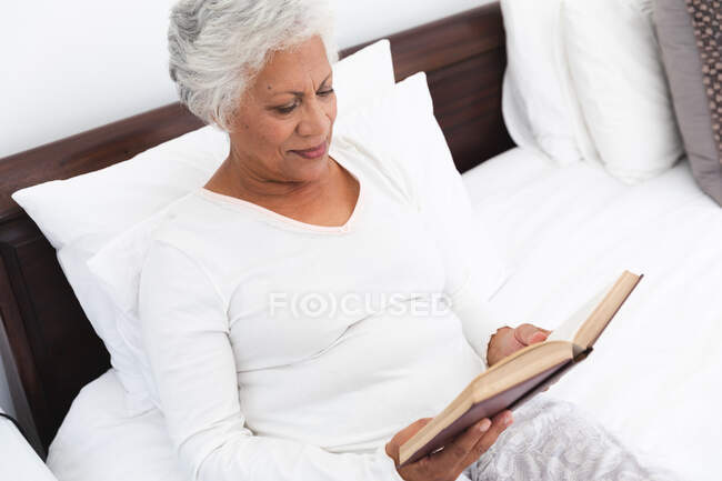 Gros plan d'une séduisante femme afro-américaine retraitée à la maison, assise dans son lit, lisant un livre et souriant, s'isolant elle-même pendant une pandémie de coronavirus covid19 — Photo de stock