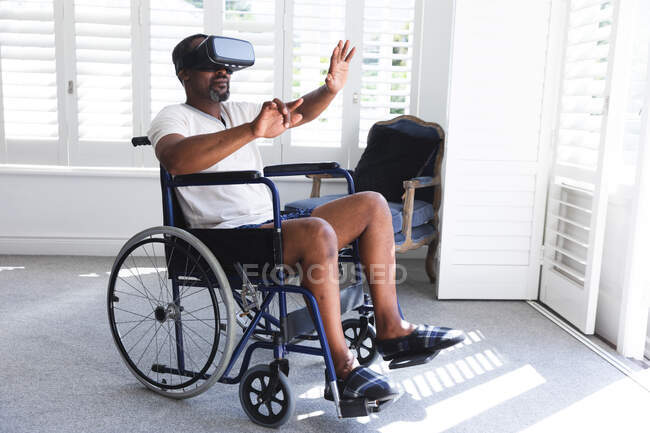 Um idoso aposentado afro-americano em casa, sentado em uma cadeira de rodas em sua roupa interior na frente de uma janela em um dia ensolarado usando um fone de ouvido VR com os braços estendidos na frente dele, auto-isolante durante a pandemia do coronavírus covid19 — Fotografia de Stock