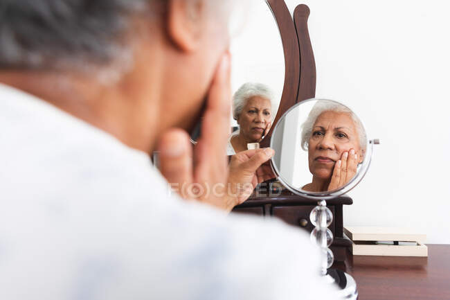 Uma mulher afro-americana aposentada sênior em casa em seu quarto, sentada em sua mesa de vestir olhando para seu reflexo no espelho e tocando seu rosto, auto-isolante durante a pandemia do coronavírus covid19 — Fotografia de Stock