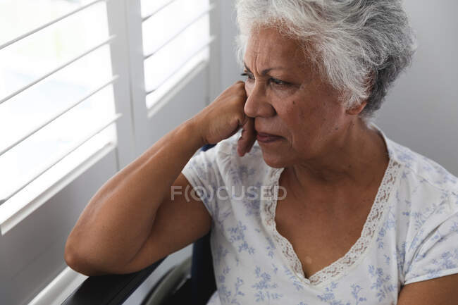 Gros plan d'une femme afro-américaine retraitée âgée à la maison, assise dans un fauteuil roulant portant un pyjama regardant par la fenêtre par une journée ensoleillée en pensée, s'isolant elle-même pendant une pandémie de coronavirus covid19 — Photo de stock