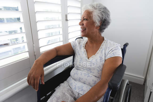 Una anciana afroamericana jubilada en casa, sentada en una silla de ruedas con pijama mirando por una ventana en un día soleado y sonriente, auto aislada durante la pandemia de coronavirus covid19 - foto de stock