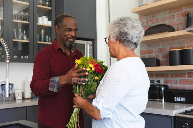 Un couple afro-américain âgé profitant de leur retraite, debout dans leur cuisine par une journée ensoleillée, l'homme tenant un bouquet de fleurs, souriant et les donnant à sa femme, à la maison ensemble isolant pendant la pandémie de coronavirus covid19 — Photo de stock