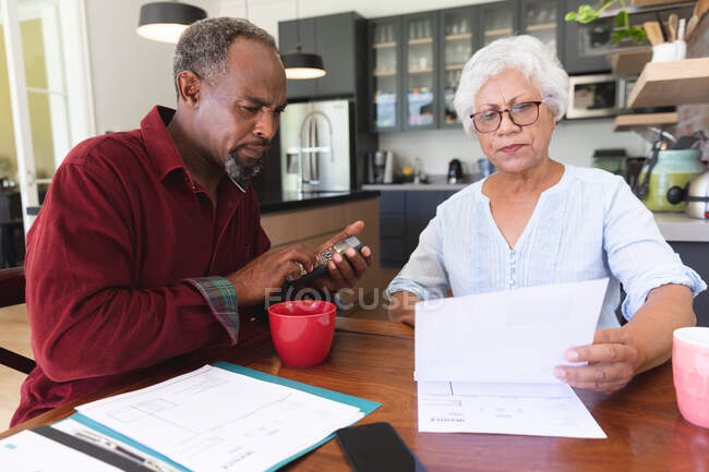Una coppia di anziani afroamericani in pensione seduti a un tavolo nella loro sala da pranzo a bere caffè, a guardare i documenti e discutere le loro finanze, a casa isolandosi insieme durante la pandemia di coronavirus19 — Foto stock