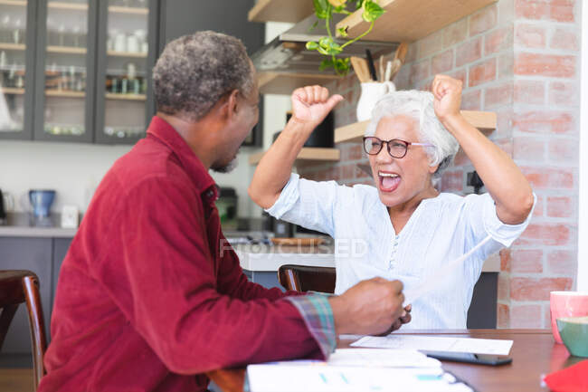 Una pareja de ancianos afroamericanos jubilados sentados en una mesa en su comedor, mirando el papeleo y discutiendo sus finanzas, la mujer emocionada, levantando las manos y vitoreando, en casa juntos aislándose durante la pandemia del coronavirus covid19 - foto de stock