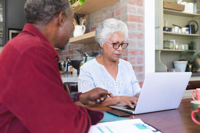 Ein älteres afroamerikanisches Rentnerehepaar sitzt an einem Tisch in ihrem Esszimmer, betrachtet Papierkram und diskutiert über ihre Finanzen, die Frau benutzt einen Laptop-Computer, zu Hause zusammen isoliert während der Coronavirus-Covid19 Pandemie — Stockfoto