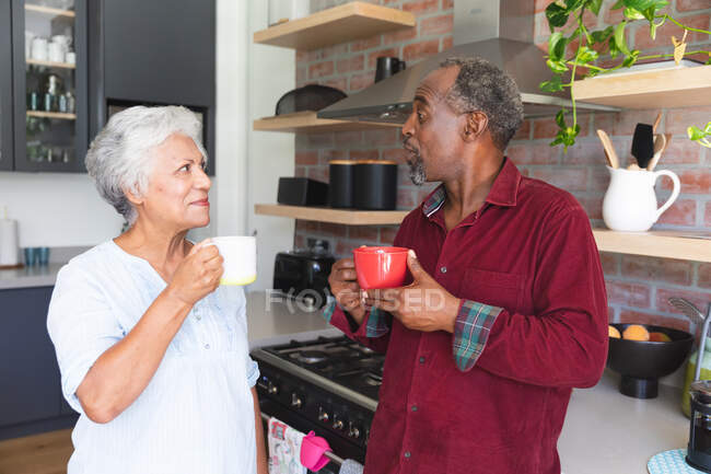 Glückliche ältere afroamerikanische Rentnerehepaar zu Hause in ihrer Küche stehen, reden und Kaffee trinken, zu Hause zusammen isolieren während Coronavirus covid19 Pandemie — Stockfoto