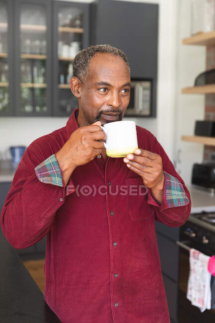 Feliz anciano jubilado afroamericano en casa de pie en la cocina, sonriendo y bebiendo una taza de café, en casa aislando durante coronavirus covid19 pandemia - foto de stock