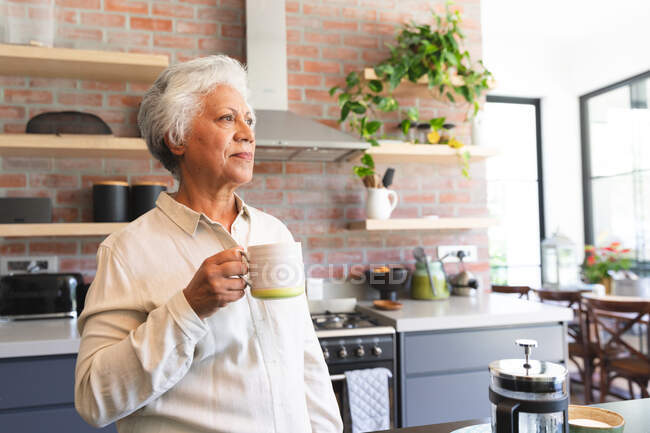 Mujer afroamericana jubilada mayor en casa parada en la cocina, sosteniendo una taza de café y mirando hacia otro lado, aislada en casa durante la pandemia de coronavirus covid19 - foto de stock