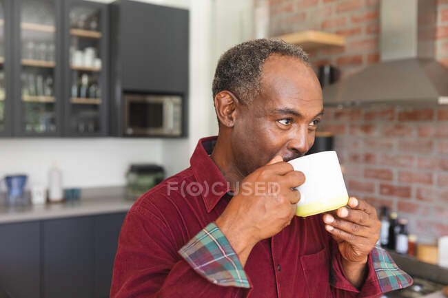 Счастливый пожилой афроамериканец в отставке дома, стоящий на кухне, улыбающийся и пьющий кружку кофе, дома изолирующий во время пандемии коронавируса covid19 — стоковое фото