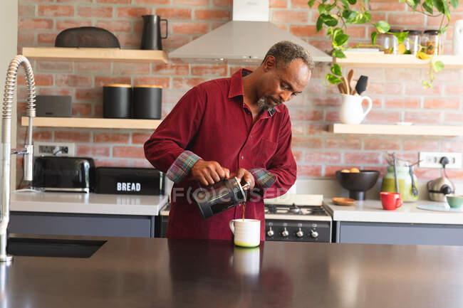 Senior uomo afroamericano in pensione a casa in piedi in cucina, versando con attenzione una tazza di caffè da un caffè, a casa isolando durante il coronavirus covid19 pandemia — Foto stock