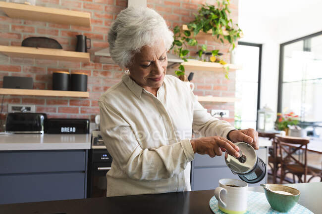 Старшая афроамериканка в отставке дома, стоящая на кухне, осторожно наливая кружку кофе из кафе, дома изолируя во время пандемии коронавируса — стоковое фото