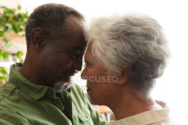 Крупным планом счастливой пожилой афроамериканской пары дома, стоящей на своей кухне, трогающей головы и смотрящей друг на друга, обнимаясь, дома вместе изолирующей во время пандемии коронавируса — стоковое фото