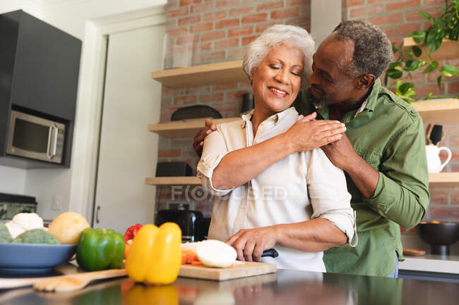 Счастливая пожилая афроамериканская пара дома, готовит еду, режет овощи и обнимает их на кухне, дома вместе изолируя во время пандемии коронавируса — стоковое фото