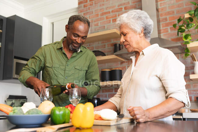 Счастливая пожилая афроамериканская пара дома, готовит овощи, чтобы приготовить еду, и мужчина наливает им бокалы вина, пара дома вместе изолирует во время пандемии коронавируса — стоковое фото