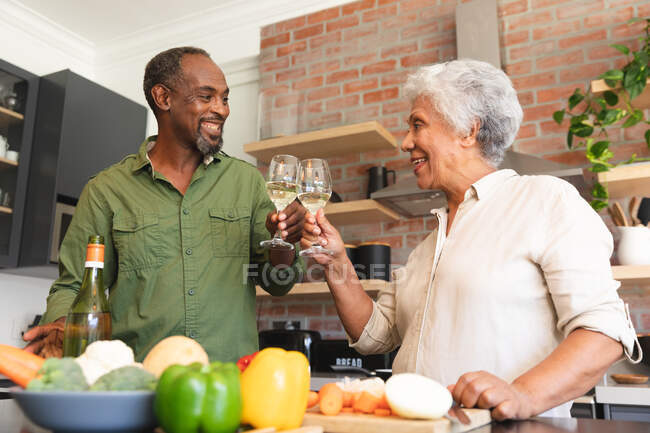 Happy senior aposentado casal afro-americano em casa, preparando legumes para fazer uma refeição, e fazer um brinde com copos de vinho branco, casal em casa juntos isolando durante coronavírus covid19 pandemia — Fotografia de Stock
