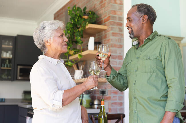 Feliz pareja jubilada afroamericana jubilada en casa, sonriendo y haciendo un brindis con copas de vino blanco levantadas, pareja en casa aislándose durante la pandemia de coronavirus covid19 - foto de stock