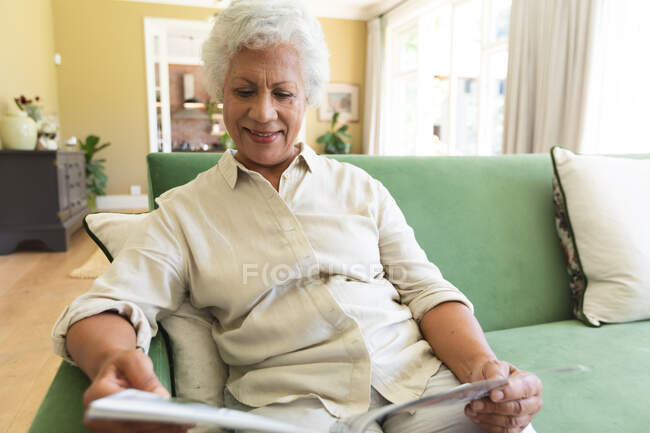 Primer plano de feliz anciano jubilado mujer afroamericana en casa sentada en su sala de estar, leyendo una revista y sonriendo, auto aislamiento durante coronavirus covid19 pandemia - foto de stock