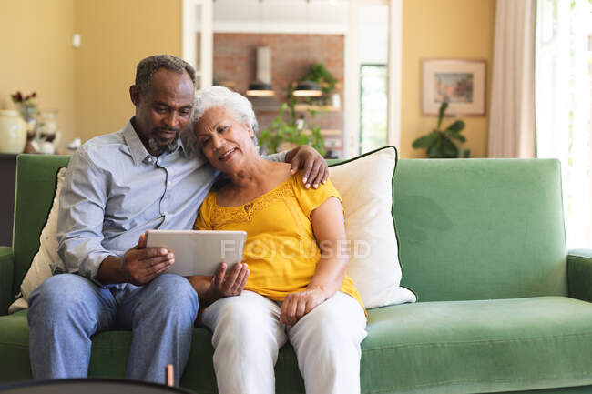 Felice anziano pensionato coppia afroamericana a casa seduta su un divano nel loro soggiorno, abbracciando e utilizzando un computer tablet insieme e sorridente, coppia isolante durante coronavirus covid19 pandemia — Foto stock