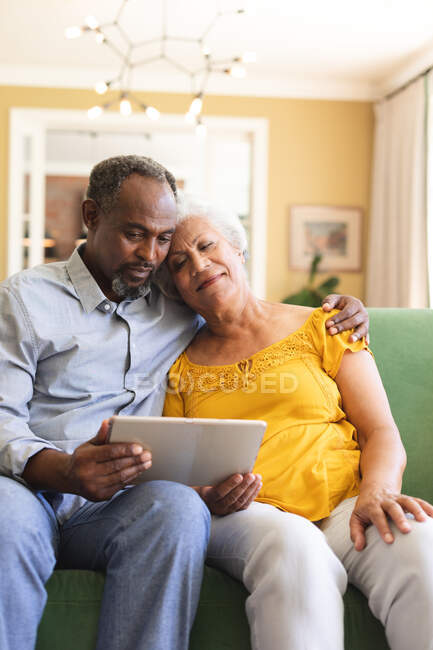 Зблизька щаслива подружня пара афроамериканців у відставці, яка сидить вдома на дивані у своїй вітальні, обіймає і використовує планшетний комп'ютер разом і посміхається, пара ізолюється під час коронавірусної ковірусної ковідемії — стокове фото