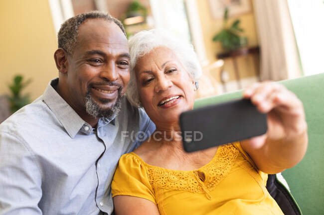Закрийте щасливу американську пару на пенсії вдома в своїй вітальні, сидячи на дивані, жінка тримає смартфон, дивлячись на телефон разом, беручи селфі і посміхаючись — стокове фото