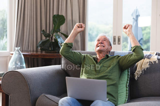 Um homem branco aposentado bonito feliz aposentado em casa sentado em um sofá em sua sala de estar, usando um computador portátil, sorrindo e acenando com os punhos no ar, celebrando um sucesso, auto-isolando durante a pandemia do coronavírus covid19 — Fotografia de Stock