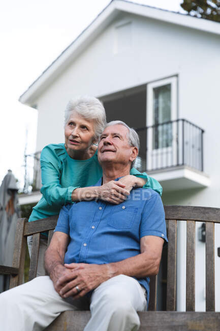 Großaufnahme eines glücklichen Rentnerehepaares aus dem Kaukasus zu Hause im Garten vor ihrem Haus, der Mann sitzt auf einer Bank und die Frau hinter ihm umarmt ihn, beide schauen weg und lächeln — Stockfoto