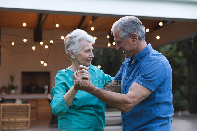 Счастливая пожилая кавказская пара дома, держась за руки, танцуя вместе в саду и улыбаясь, дома вместе изолируют во время пандемии коронавируса — стоковое фото