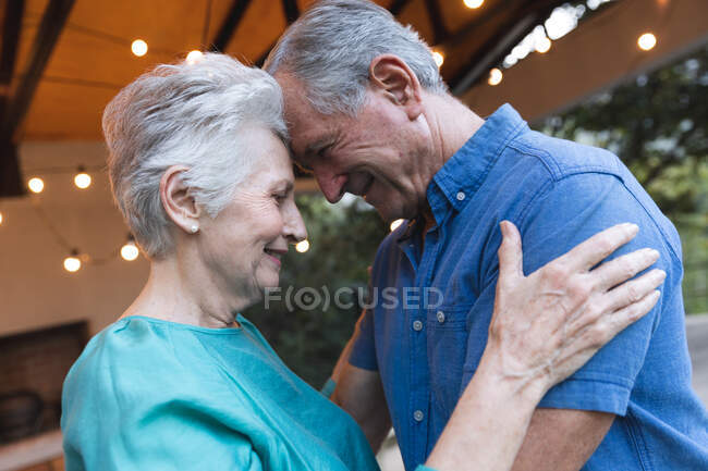 Vista lateral de una feliz pareja de ancianos caucásicos jubilados en casa, tocando cabezas juntos y mirándose mientras abrazan, en casa juntos aislando durante coronavirus covid19 pandemia - foto de stock