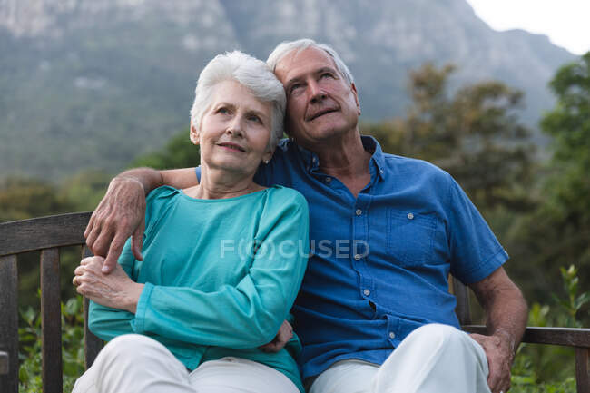 Heureux couple de personnes âgées caucasiennes à la retraite à la maison dans le jardin à l'extérieur de leur maison, assis sur un banc, relaxant et embrassant, les deux regardant loin et souriant, à la maison ensemble isolant pendant la pandémie de coronavirus covid19 — Photo de stock