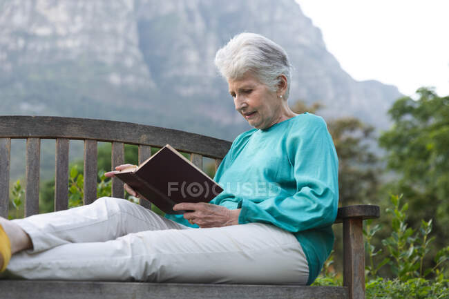 Счастливая старшая белая женщина на пенсии дома в саду снаружи, сидя на скамейке, читая книгу с поднятыми ногами, расслабляясь на природе, самоизоляция во время пандемии коронавируса — стоковое фото