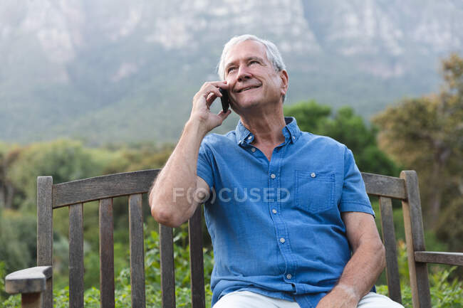 Feliz jubilado anciano caucásico en casa en el jardín fuera de su casa, sentado en un banco, relajándose en la naturaleza y hablando en un teléfono inteligente, mirando hacia otro lado y sonriendo, auto aislado durante coronavirus covid19 pandemia - foto de stock