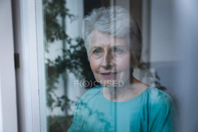 Счастливая старшая белая женщина в отставке дома смотрит в окно с отражением сада, самоизоляция во время коронавируса covid19 пандемии — стоковое фото