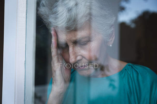 Літня жінка з Кавказу вдома стоїть біля вікна з головним болем, тримаючи голову, з відзеркаленням саду у вікні, самоізолюючись під час коронавірусу covid19 пандемії — стокове фото