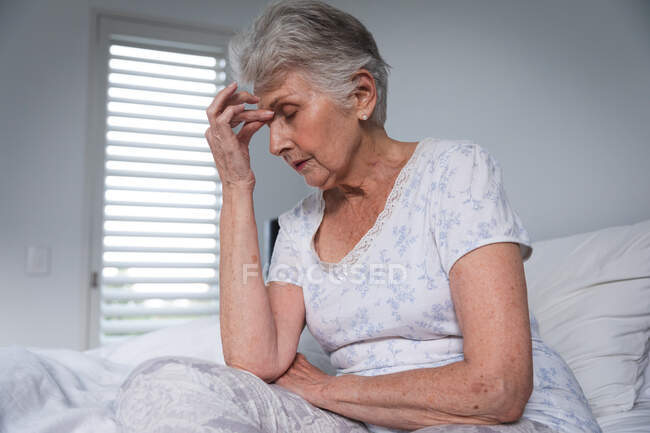 Крупным планом пожилой белой женщины в отставке, сидящей дома в постели с головной болью, держащей голову с закрытыми глазами, самоизолирующейся во время пандемии коронавируса — стоковое фото