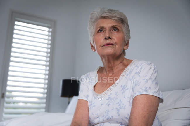 Крупным планом пожилой белой женщины в отставке, сидящей дома в постели в ночной одежде и отводящей взгляд, изолирующей себя во время пандемии коронавируса — стоковое фото