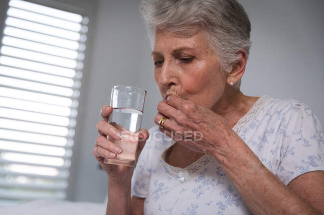 Primo piano di una donna anziana caucasica in pensione a casa seduta a letto nella sua camera da letto con in mano un bicchiere d'acqua e prendendo farmaci, auto-isolante durante la pandemia di coronavirus19 — Foto stock