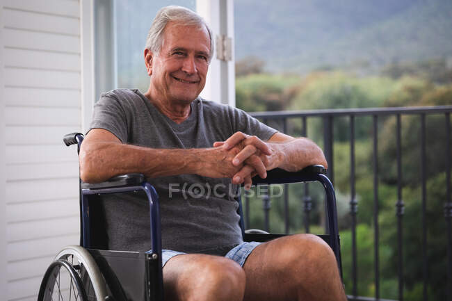 Портрет отставного старшего кавказца дома, в нижнем белье, сидящего в инвалидном кресле перед окном, в солнечный день смотрящего в камеру и улыбающегося, самоизолирующегося во время пандемии коронавируса — стоковое фото