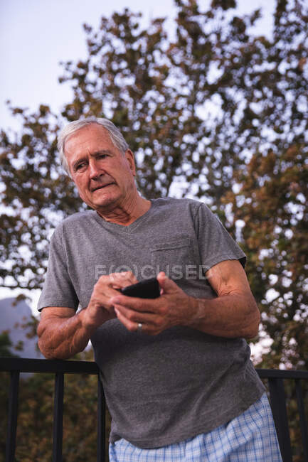 Крупный план красивого пожилого кавказца, наслаждающегося пенсией, в саду на солнце, переписывающегося с мобильным телефоном, самоизолирующегося во время пандемии коронавируса — стоковое фото
