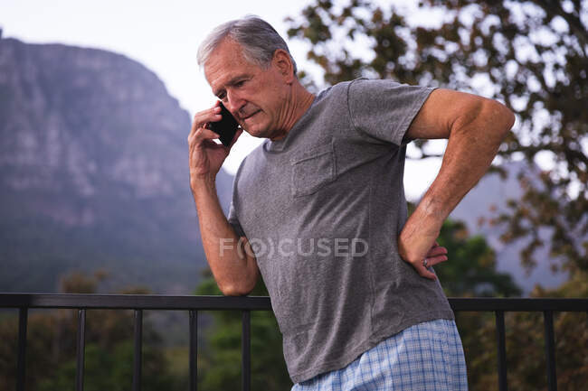 Un hombre caucásico mayor guapo disfrutando de su retiro, en un jardín al sol hablando en un teléfono móvil, auto aislándose durante la pandemia del coronavirus covid19 - foto de stock
