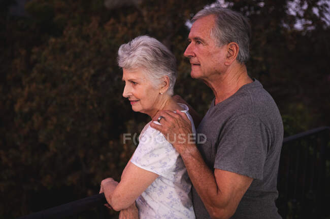 Vue latérale du couple caucasien âgé à la retraite à la maison dans leur jardin, debout ensemble, l'homme avec sa main sur l'épaule de la femme à la fois regarder loin, à la maison ensemble isolant pendant la covid19 coronavirus pandémie — Photo de stock