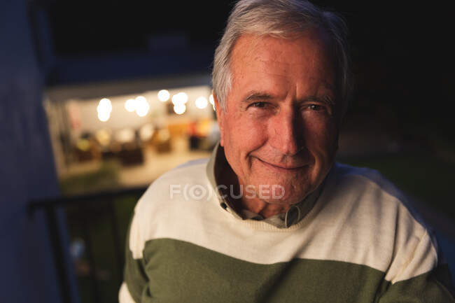 Retrato de perto de um belo homem caucasiano sênior desfrutando de sua aposentadoria, em pé fora de sua casa em uma varanda à noite olhando para longe sorrindo, auto-isolante durante coronavírus covid19 pandemia — Fotografia de Stock