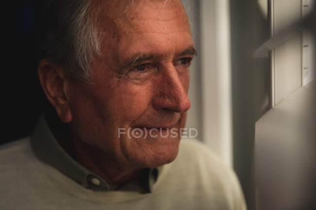 Gros plan d'un bel homme caucasien âgé profitant de sa retraite, debout à la maison, regardant par la fenêtre et souriant le soir, s'isolant lui-même pendant une pandémie de coronavirus covid19 — Photo de stock