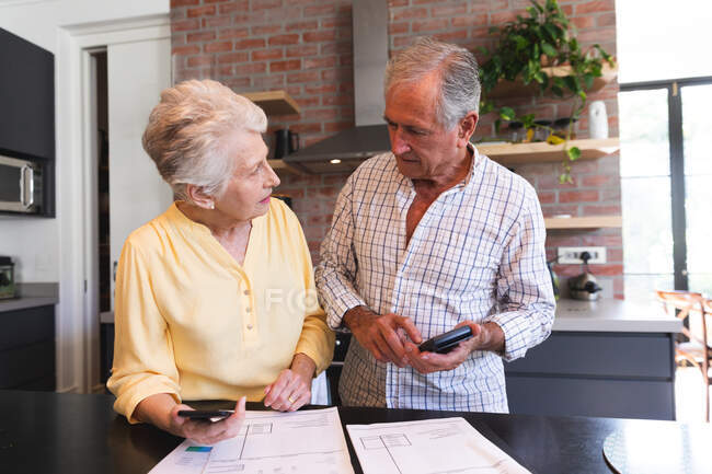A retired senior Caucasian couple standing at a table in their dining room looking at paperwork and discussing their finances, at home together isolating during coronavirus covid19 pandemic — Stock Photo
