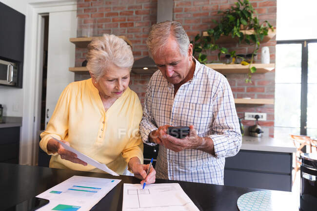 Una pareja de ancianos jubilados del Cáucaso de pie en una mesa en su comedor mirando el papeleo, usando una calculadora y discutiendo sus finanzas, en casa juntos aislando durante la pandemia de coronavirus covid19 - foto de stock