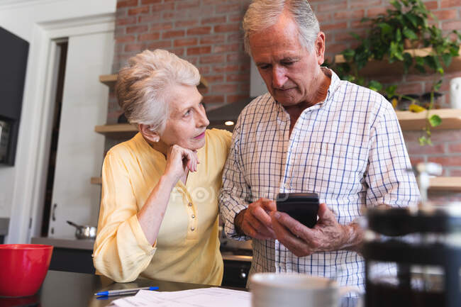 A retired senior Caucasian couple standing at a table in their dining room using a calculator and discussing their finances, with a pot of coffee and cup on the table in the foreground, at home together isolating during coronavirus covid19 pandemic — Stock Photo