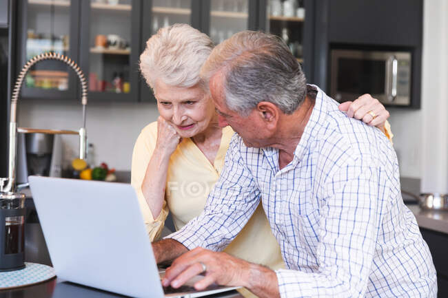 Una pareja de ancianos jubilados caucásicos en casa de pie en la encimera en su cocina, hablando y sonriendo, utilizando un ordenador portátil juntos, la mujer con el brazo alrededor del hombre, pareja aislante durante coronavirus covidemic 19 - foto de stock